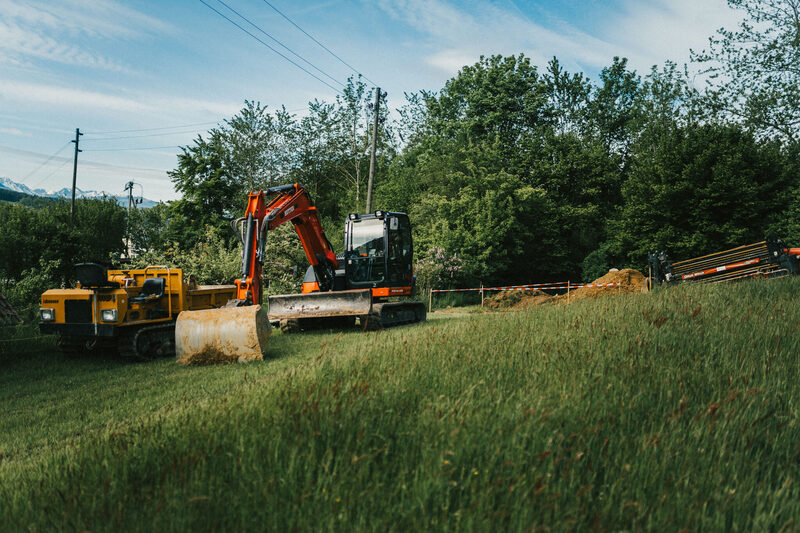 Ein grosser Raupenbagger der Firma Grabenlos AG arbeitet auf einer grünen Wiese, während im Hintergrund Bäume stehen. Die Szene verbindet Technik und Natur in harmonischer Weise.