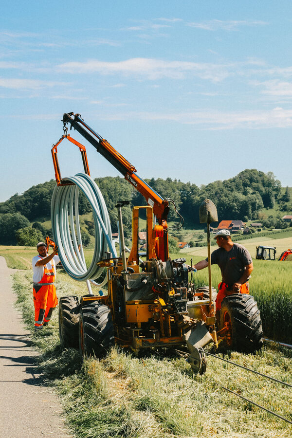 Mitarbeiter der Firma Zemp Leitungsbau GmbH stehen neben einer Pflugmaschine und hebt Rohre zum verlegen im Erdreich