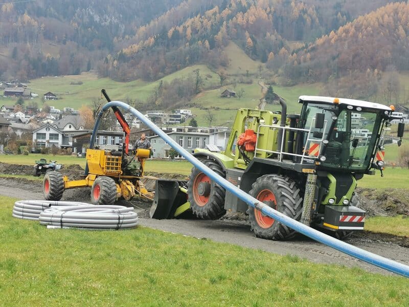 Die Pflugmaschine und ein Traktor sind geparkt, daneben liegen Kabelrohre bereit zur Verlegung. Beide Maschinen sind für den Einsatz vorbereitet, während die Rohre auf ihren Einbau warten.