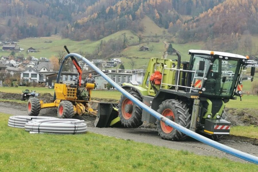 Die Pflugmaschine und ein Traktor sind geparkt, daneben liegen Kabelrohre bereit zur Verlegung. Beide Maschinen sind für den Einsatz vorbereitet, während die Rohre auf ihren Einbau warten.